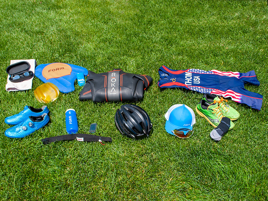 Various triathlon gear laid out on the grass including Form swim goggles, water bottle, swim cap, a helmet, and running shoes
