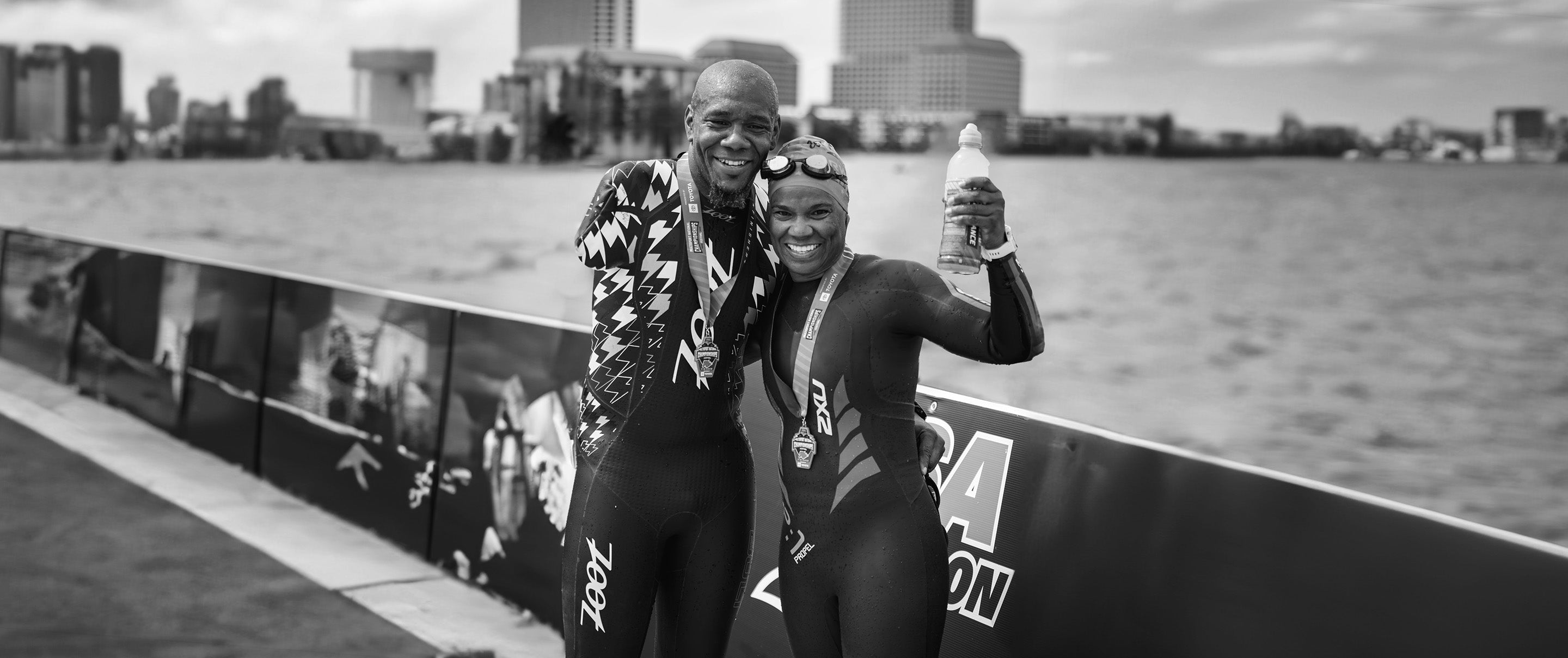 Man and woman wearing wetsuits and medals standing on a bridge